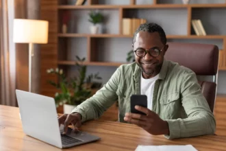 African man involved in gig economy looking at his phone and working on his computer